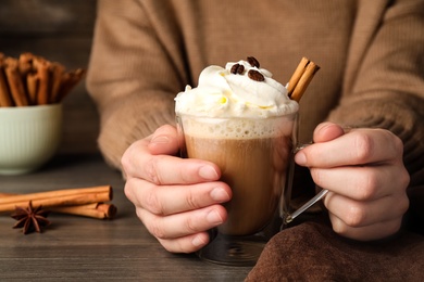 Photo of Woman holding cup of delicious coffee with whipped cream and cinnamon at wooden table, closeup