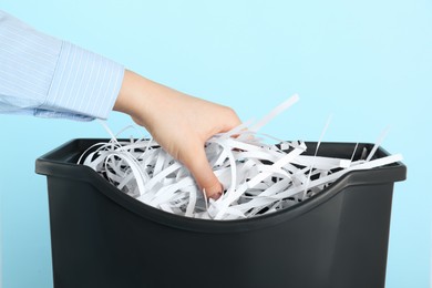 Woman putting shredded paper strips into trash bin on light blue background, closeup