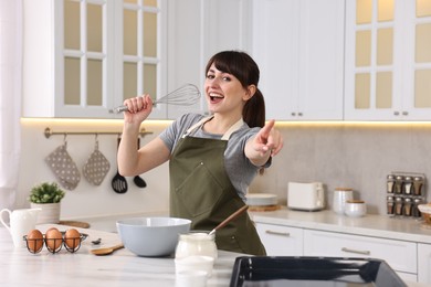 Happy young housewife with whisk having fun while cooking at white marble table in kitchen