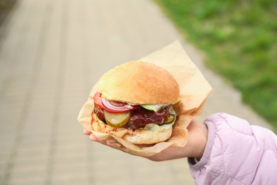 Photo of Little girl holding fresh delicious burger outdoors, closeup. Street food