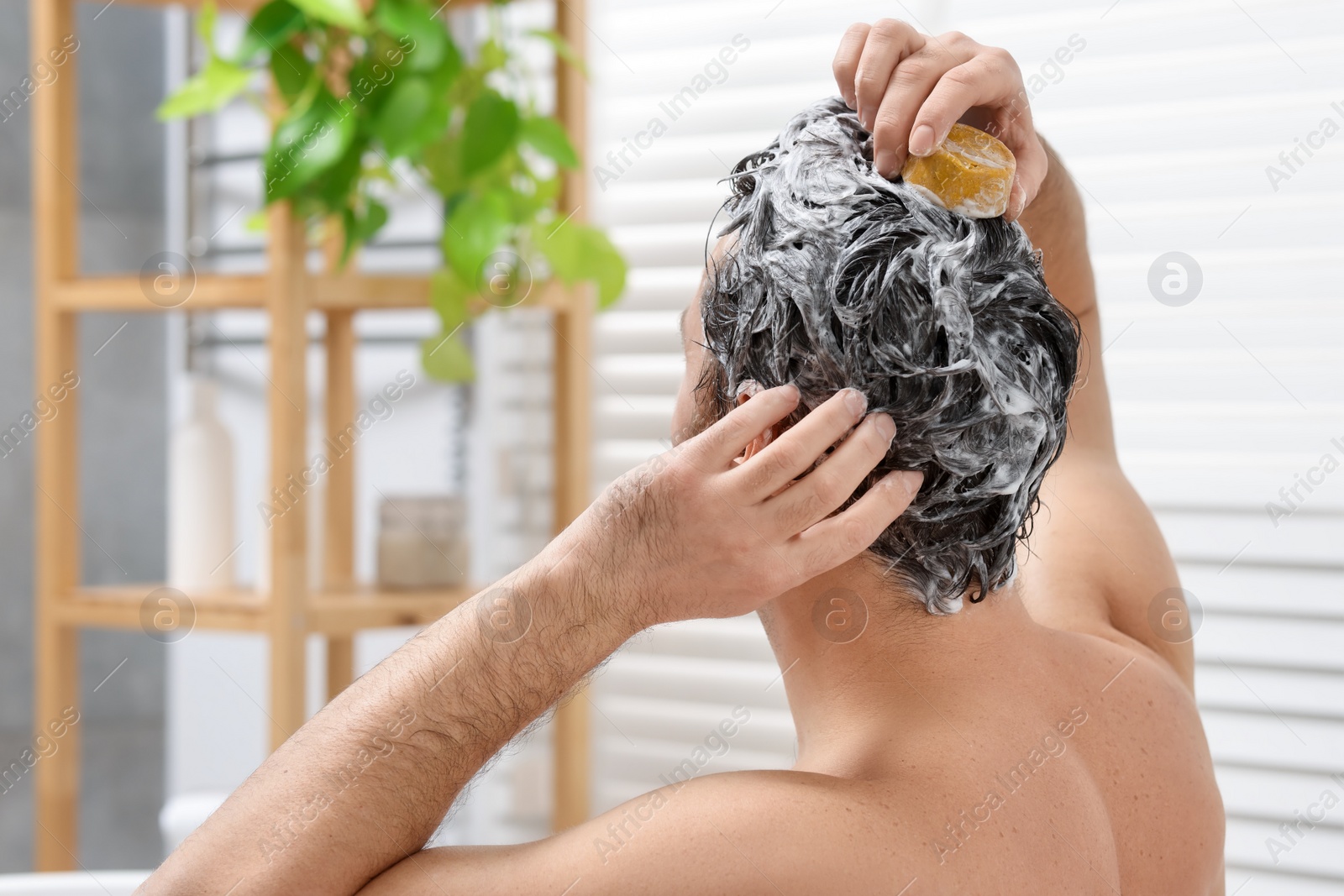 Photo of Man washing his hair with solid shampoo bar in bathroom, back view. Space for text