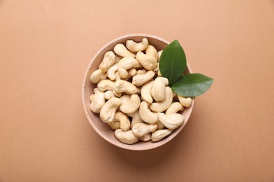 Photo of Tasty cashew nuts and green leaves in bowl on pale brown background, top view