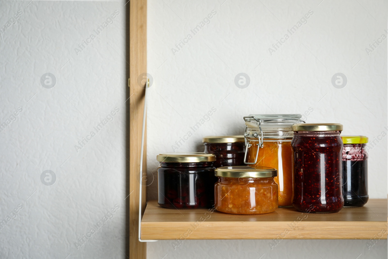 Photo of Jars with different homemade preserves on wooden shelf near white wall