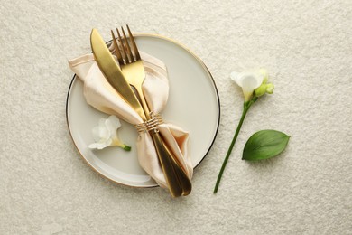 Stylish setting with cutlery, napkin, flowers and plate on light textured table, flat lay