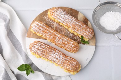 Photo of Delicious eclairs filled with cream and powdered sugar on white tiled table, top view