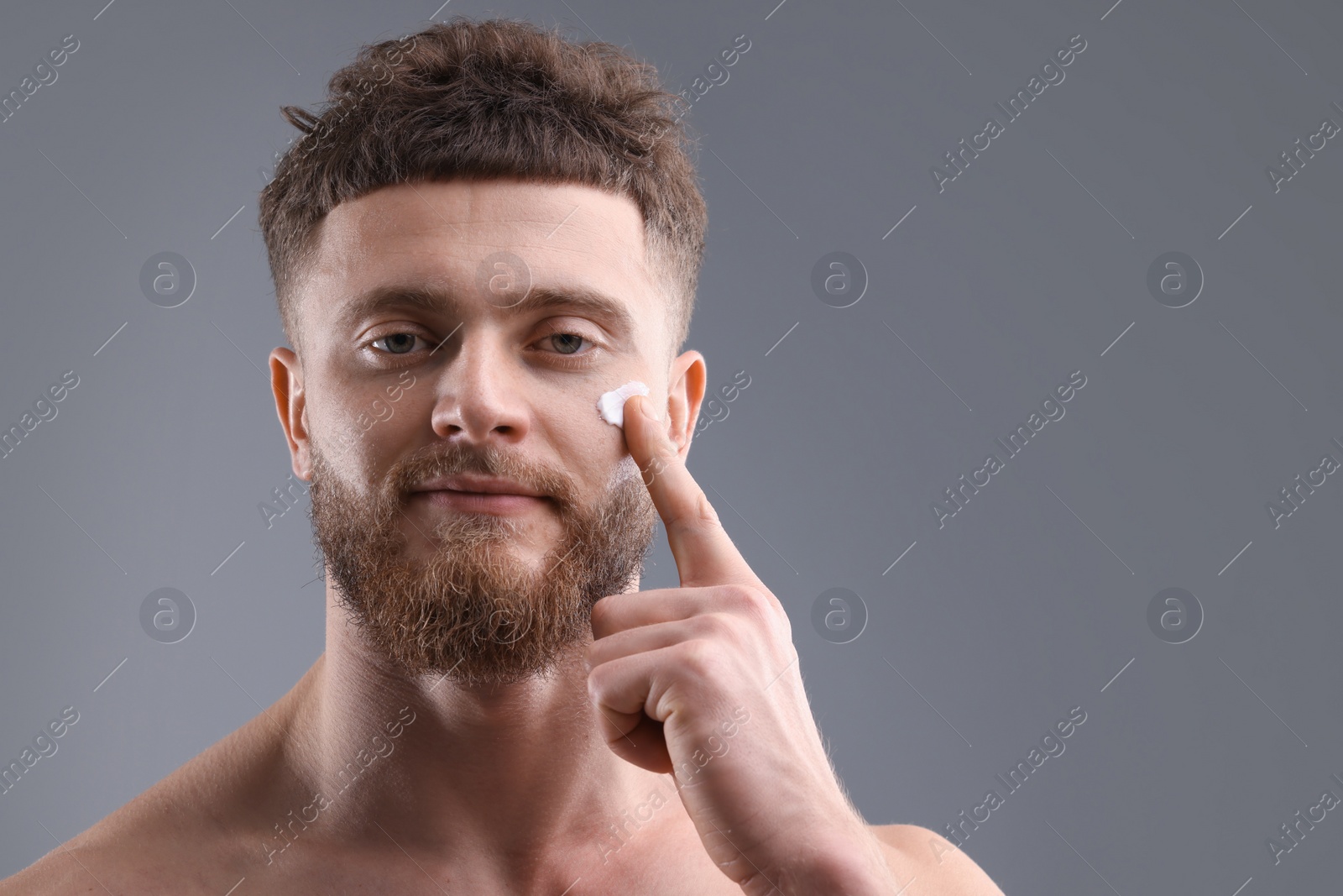 Photo of Handsome man applying moisturizing cream onto his face on grey background, space for text