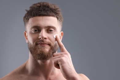 Handsome man applying moisturizing cream onto his face on grey background, space for text