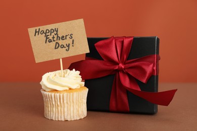 Photo of Happy Father's Day. Tasty cupcake with greetings and gift box on brown table, closeup