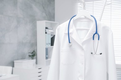 White doctor's gown and stethoscope hanging on rack in clinic, closeup. Space for text