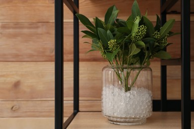 White filler with green branches in glass vase on shelf against wooden wall. Water beads