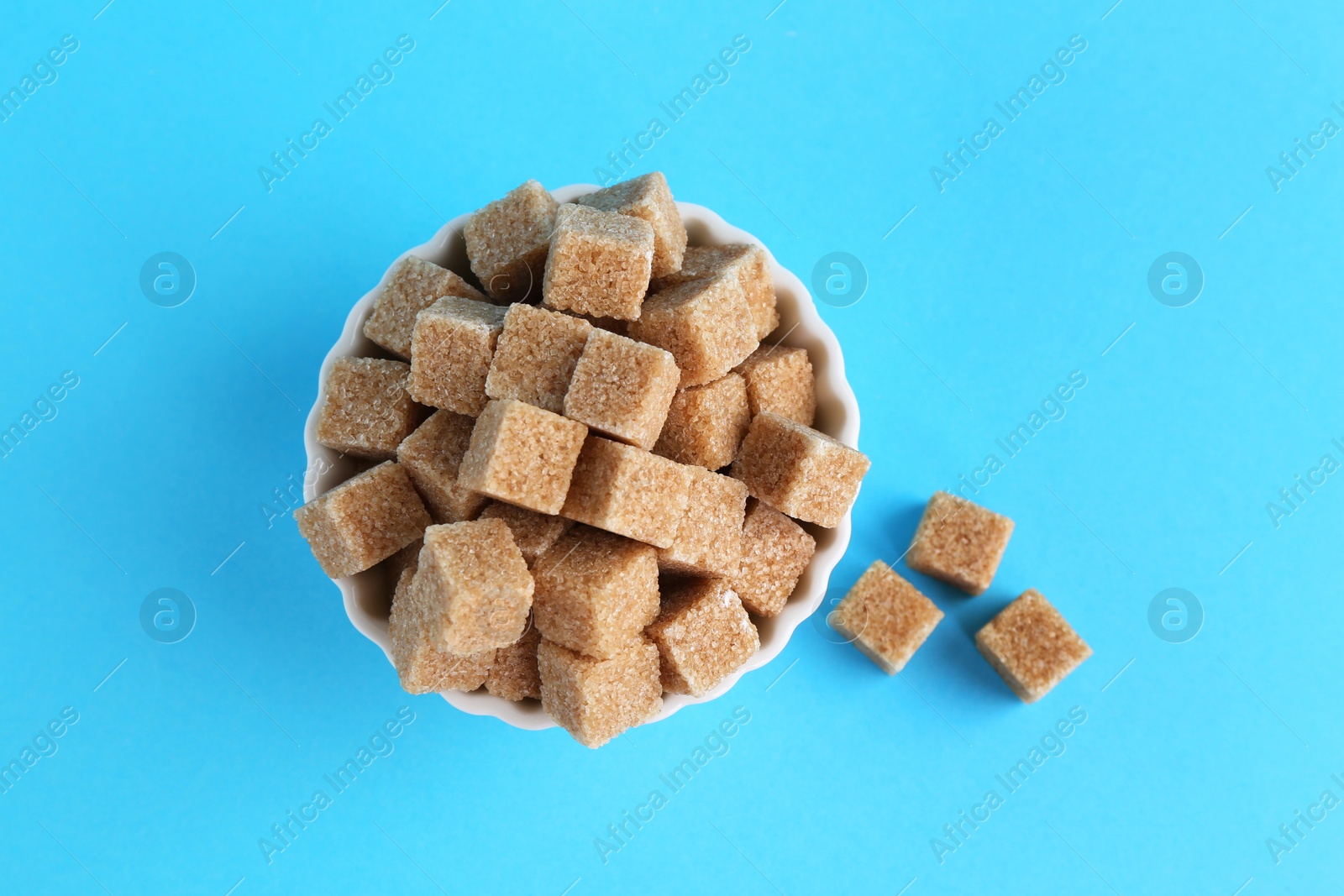 Photo of Brown sugar cubes in bowl on light blue background, top view