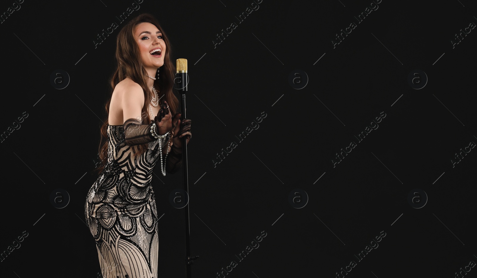 Photo of Beautiful young woman in stylish dress with microphone singing on black background, space for text