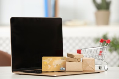 Online payment concept. Small shopping cart with bank card, boxes and laptop on white table