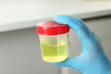 Photo of Doctor wearing glove holding container with urine sample for analysis indoors, closeup