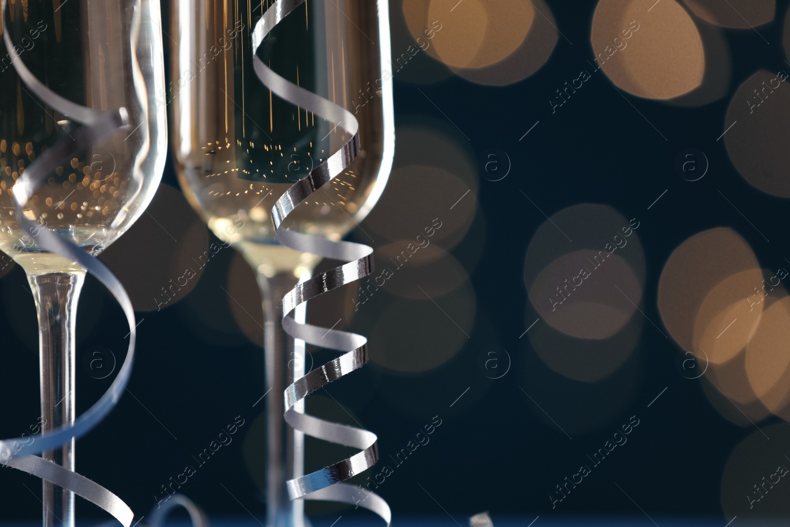 Photo of Glasses of champagne and serpentine streamers against dark blue background with blurred lights, closeup. Space for text