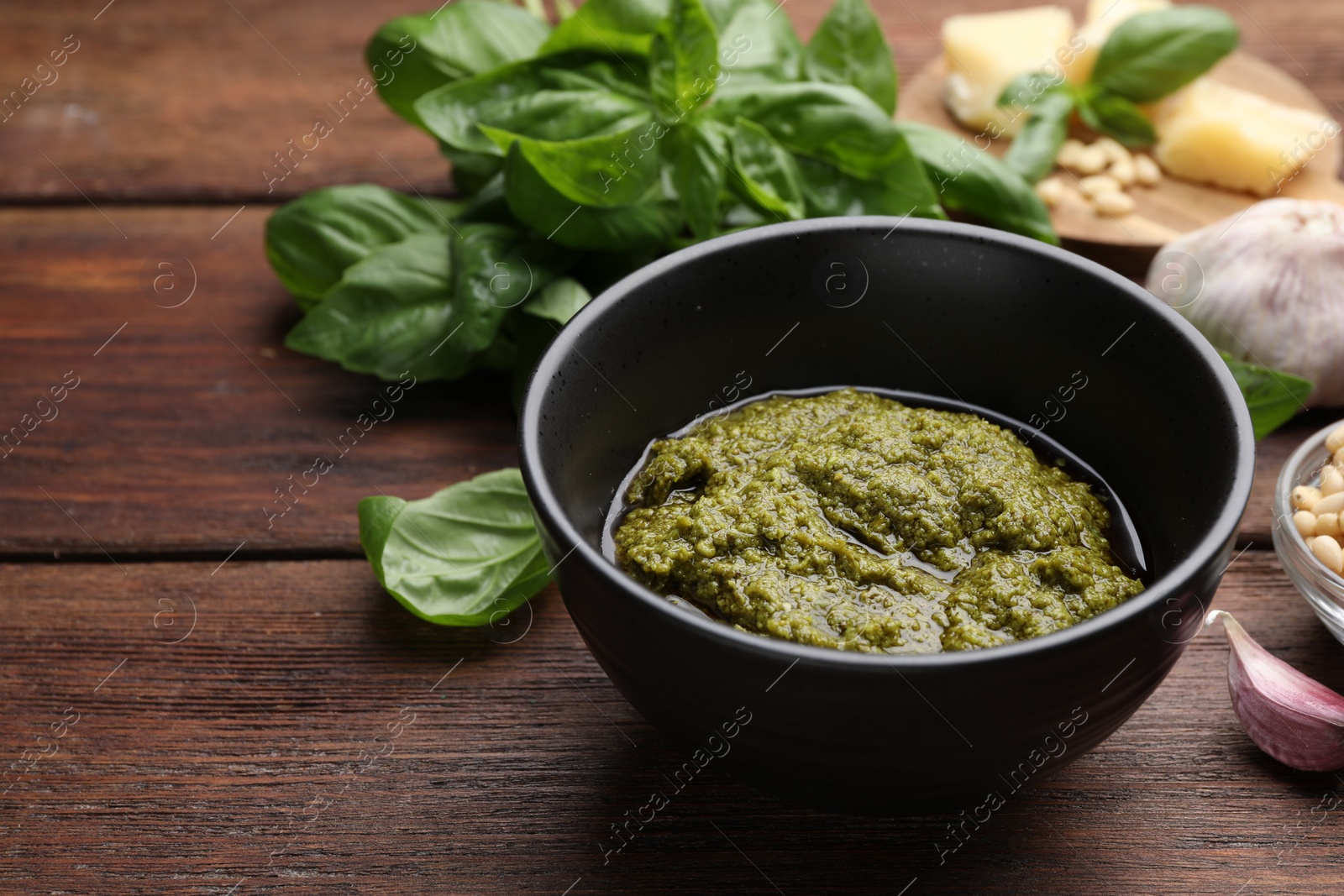 Photo of Tasty pesto sauce in bowl, basil, garlic and pine nuts on wooden table, closeup