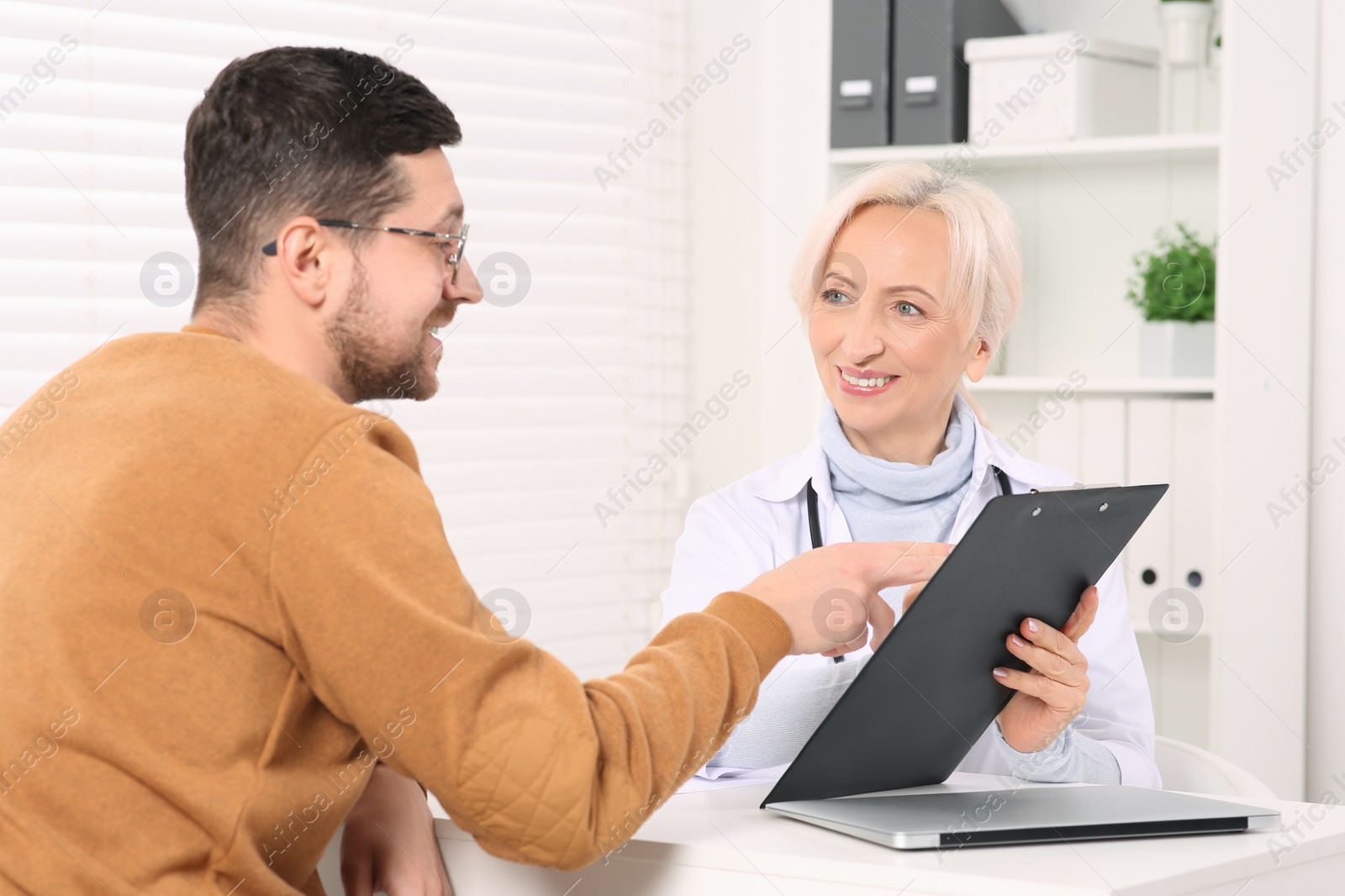 Photo of Doctor with clipboard consulting patient at white table in clinic