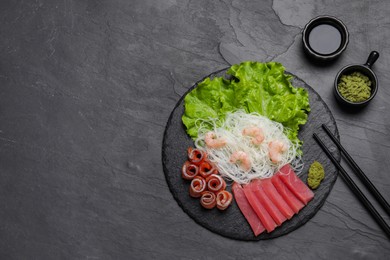 Photo of Sashimi set (raw tuna, salmon slices and shrimps) served with funchosa, lettuce, vasabi and soy sauce on dark table, flat lay. Space for text