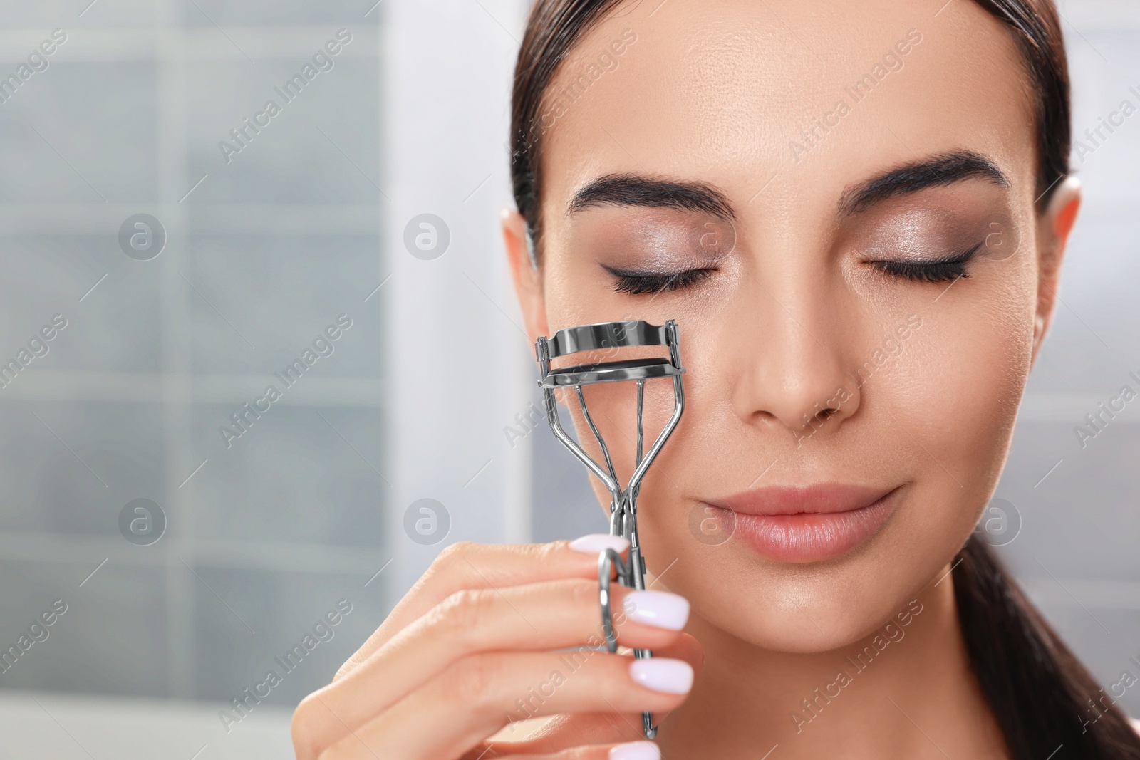 Photo of Beautiful young woman using eyelash curler in bathroom, closeup. Space for text