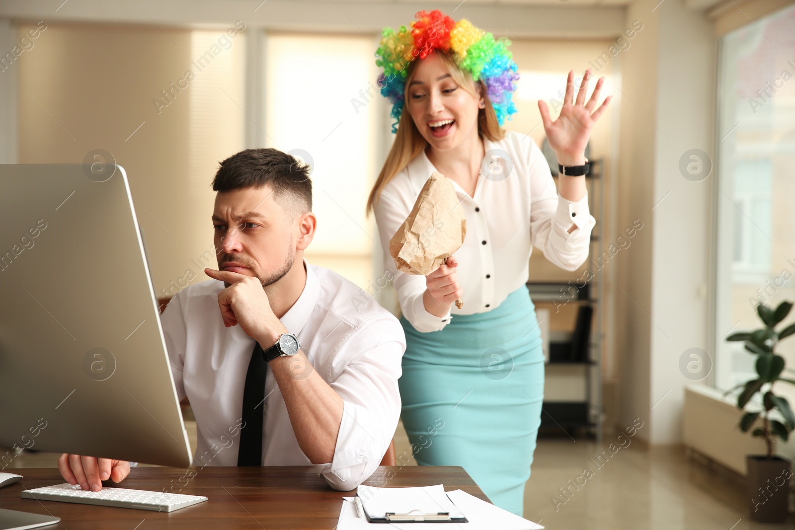 Photo of Young woman popping paper bag behind her colleague in office. Funny joke