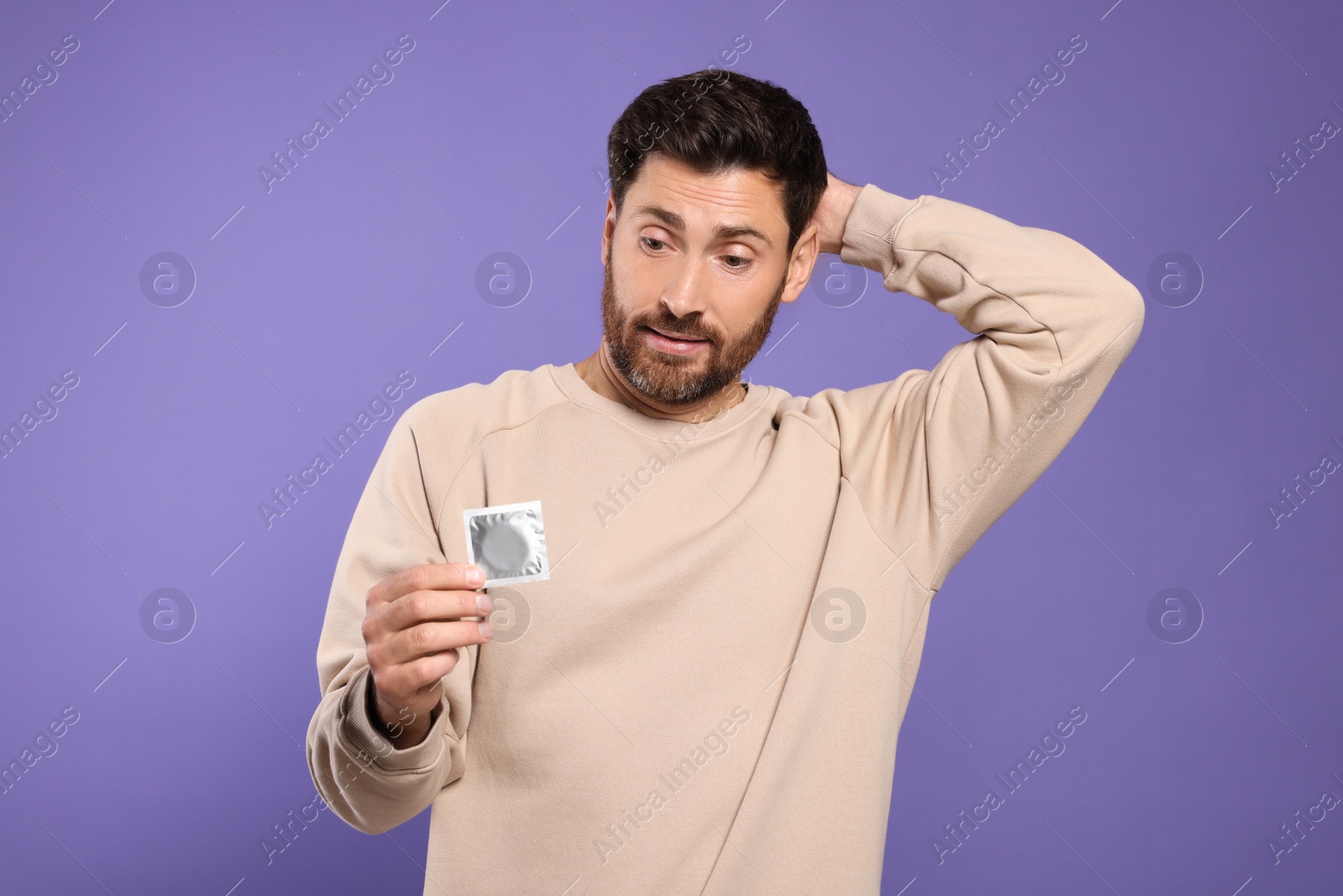 Photo of Confused man holding condom on purple background. Safe sex