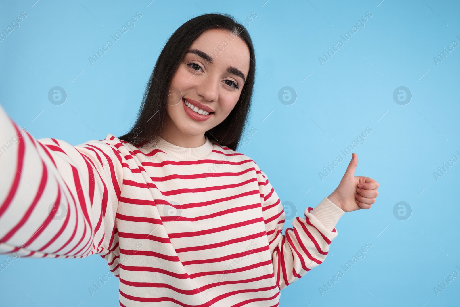 Photo of Smiling young woman taking selfie and showing thumbs up on light blue background