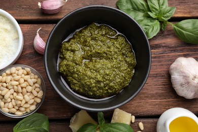 Photo of Tasty pesto sauce in bowl, basil, pine nuts, cheese, garlic and oil on wooden table, flat lay