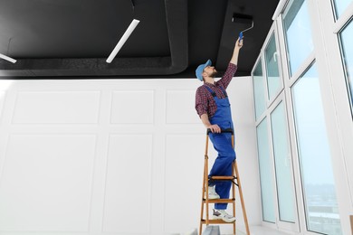 Worker in uniform painting ceiling with roller on stepladder indoors. Space for text