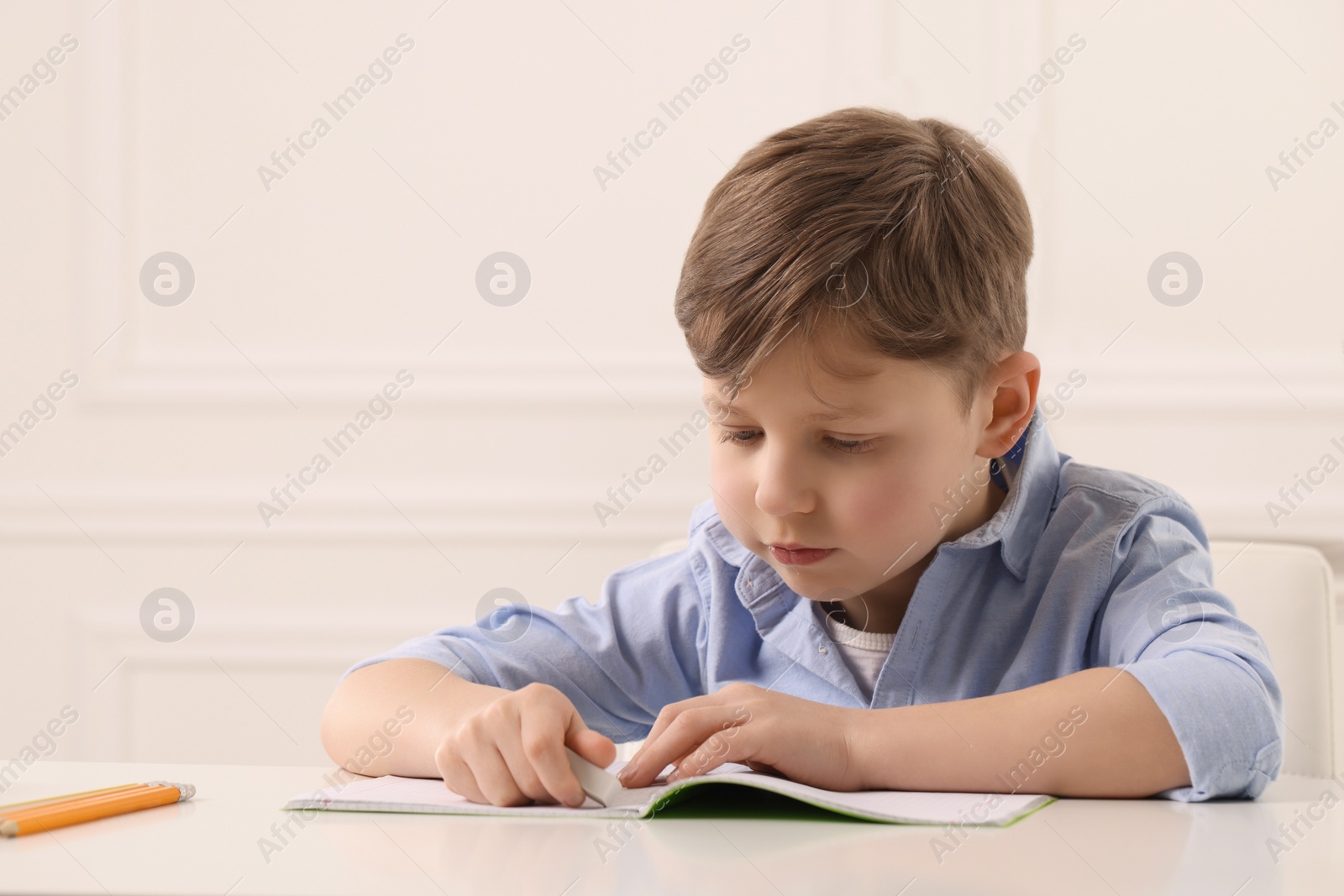 Photo of Little boy erasing mistake in his notebook at white desk indoors