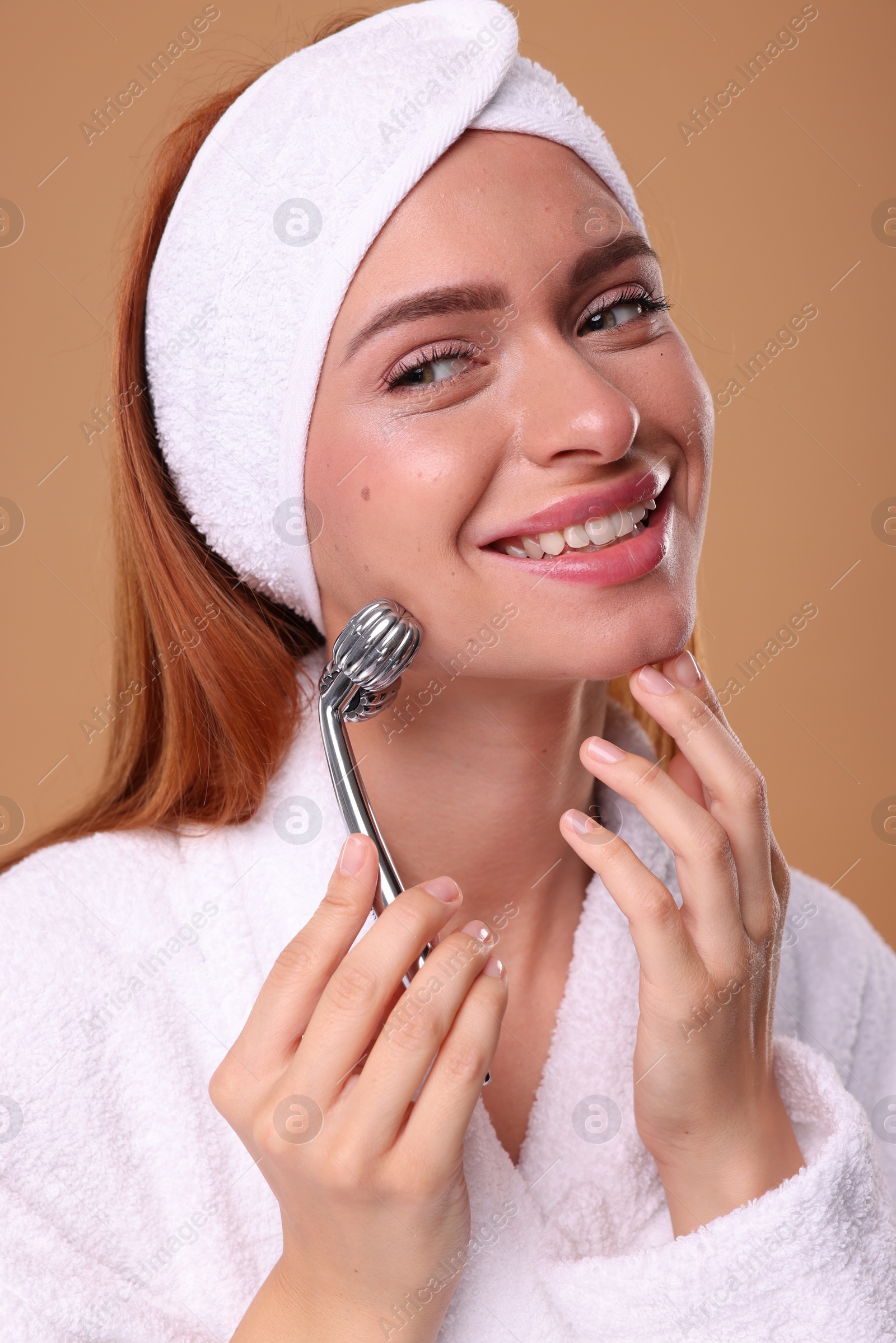 Photo of Young woman massaging her face with metal roller on pale orange background