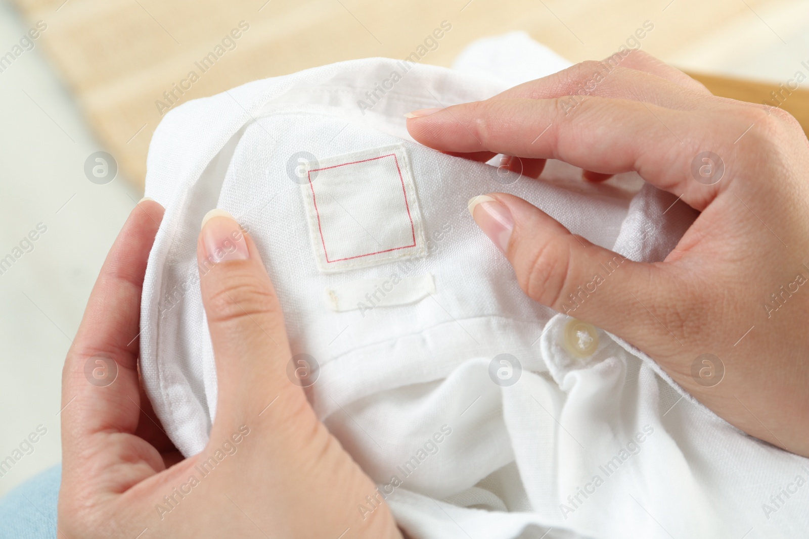 Photo of Woman holding white shirt with blank clothing label indoors, closeup