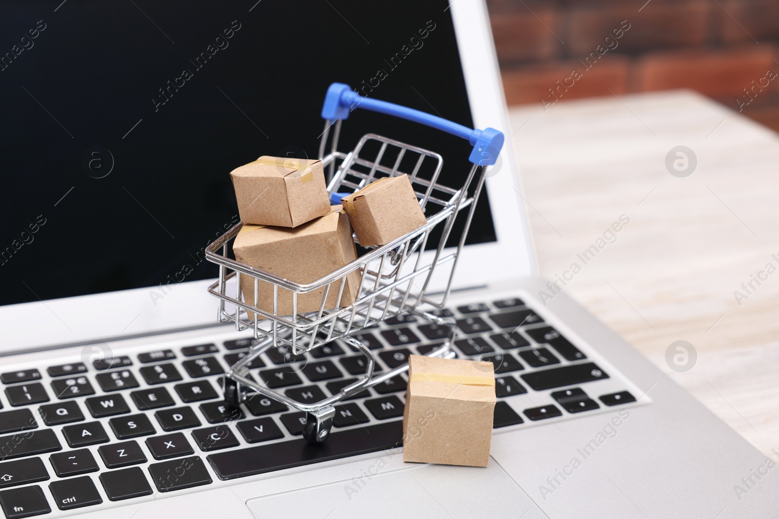 Photo of Internet store. Small cardboard boxes, shopping cart and laptop on light wooden table, closeup