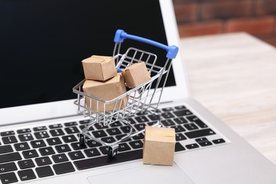 Photo of Internet store. Small cardboard boxes, shopping cart and laptop on light wooden table, closeup