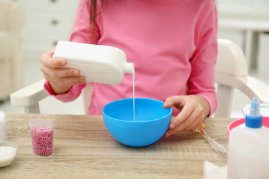 Photo of Little girl pouring glue into bowl at table in room, closeup. DIY slime toy