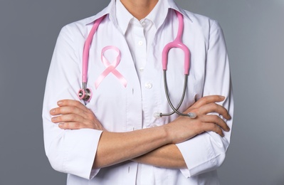 Female doctor with pink ribbon and stethoscope on grey background, closeup. Breast cancer concept