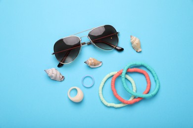 Stylish sunglasses, bracelets, seashells and rings on light blue background, flat lay