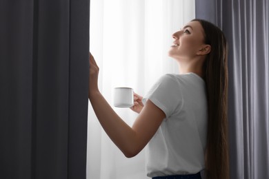 Photo of Woman with cup of hot drink opening stylish curtains at home