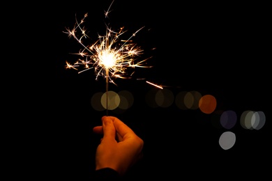 Photo of Woman holding bright burning sparklers against blurred lights, closeup. Space for text