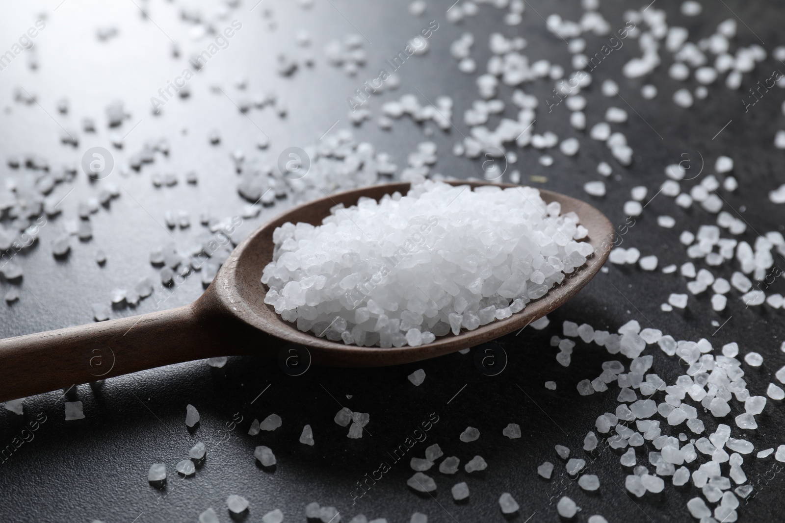 Photo of Natural salt and wooden spoon on black table, closeup