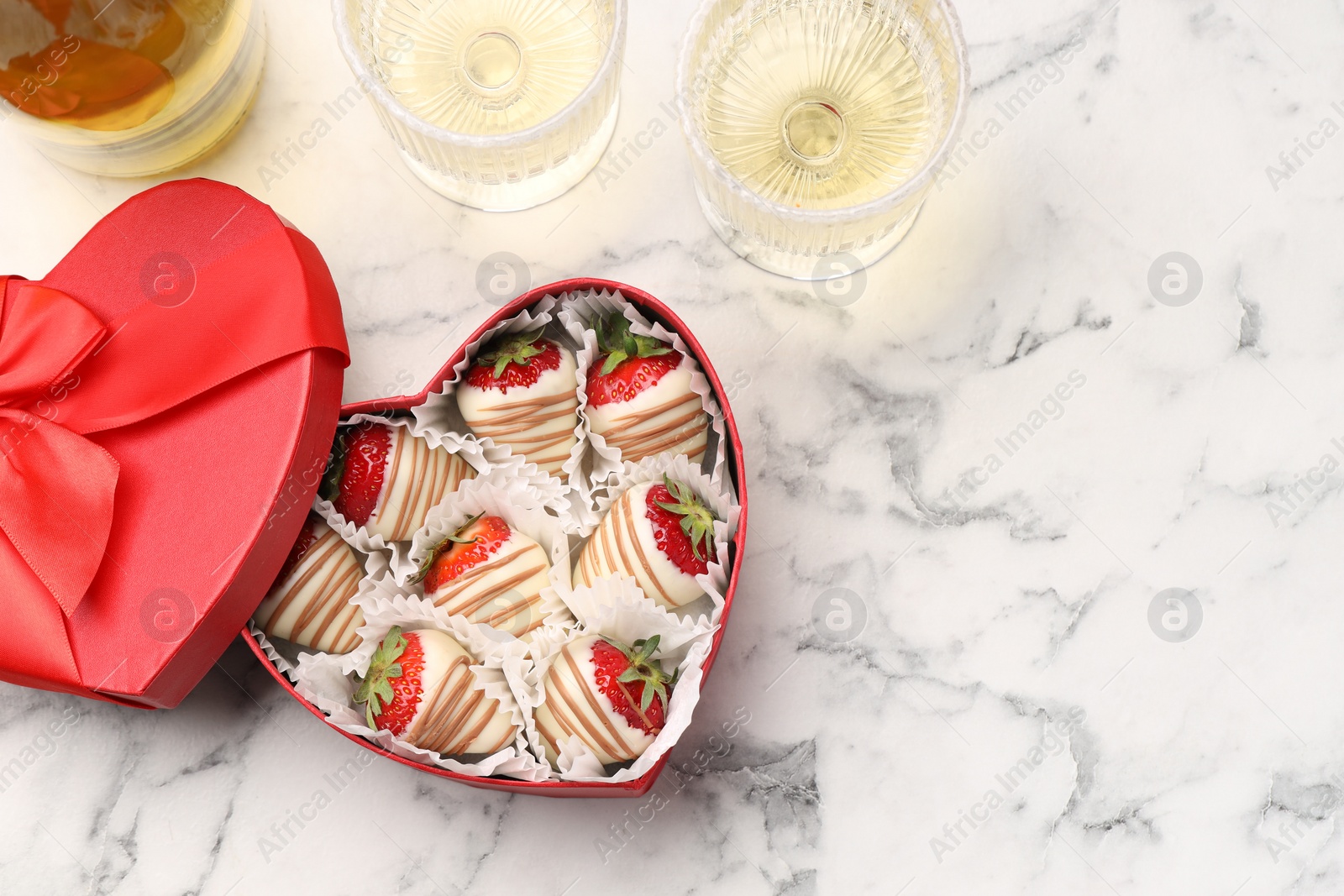 Photo of Delicious chocolate covered strawberries in heart shaped box and sparkling wine on white marble table, flat lay. Space for text