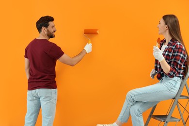 Photo of Man painting orange wall and woman sitting on folding ladder. Interior design