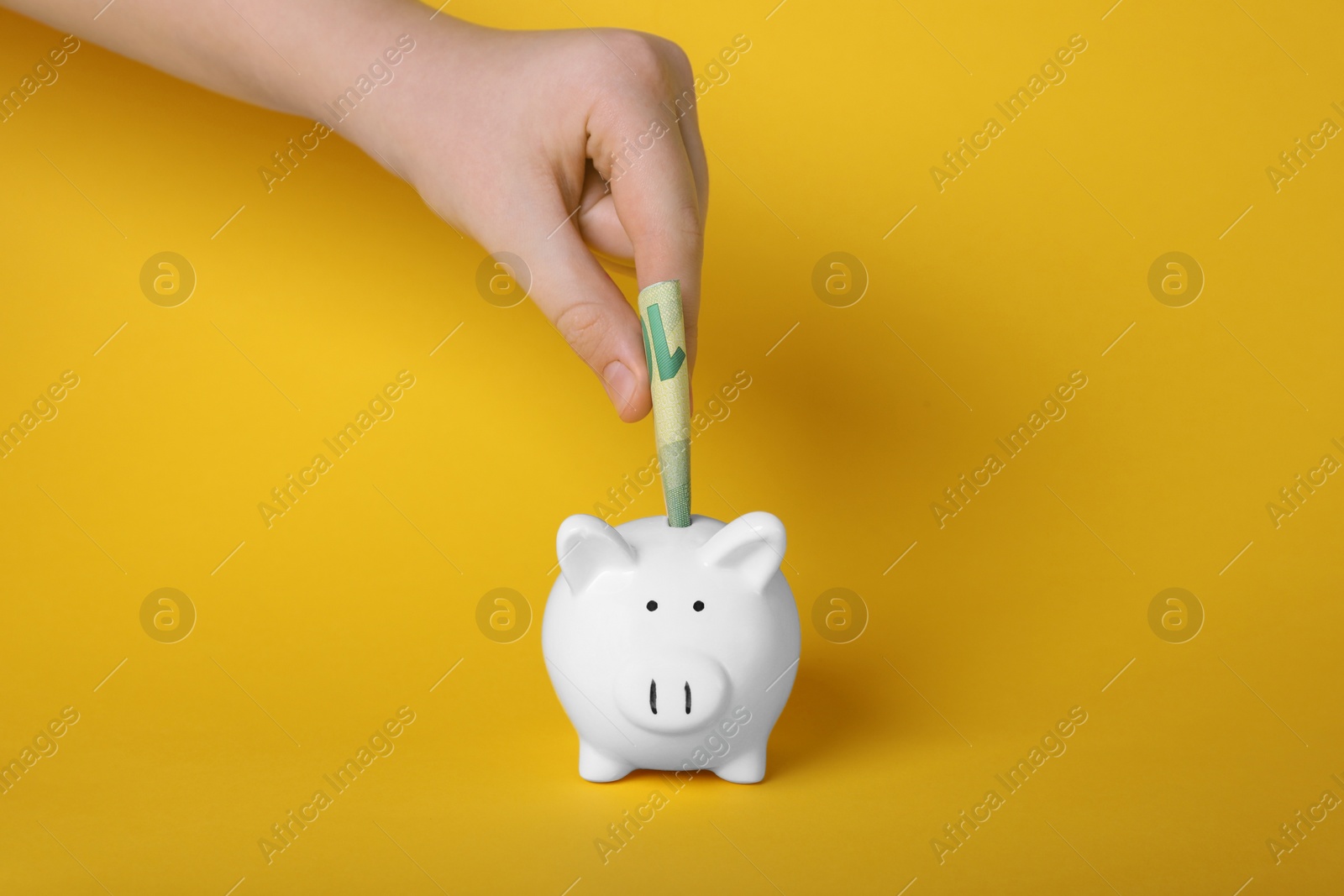Photo of Woman putting euro banknote into ceramic piggy bank on yellow background, closeup. Financial savings