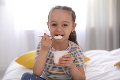 Photo of Cute little girl eating tasty yogurt on bed at home