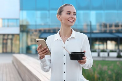 Photo of Smiling businesswoman with smartphone and lunch box outdoors