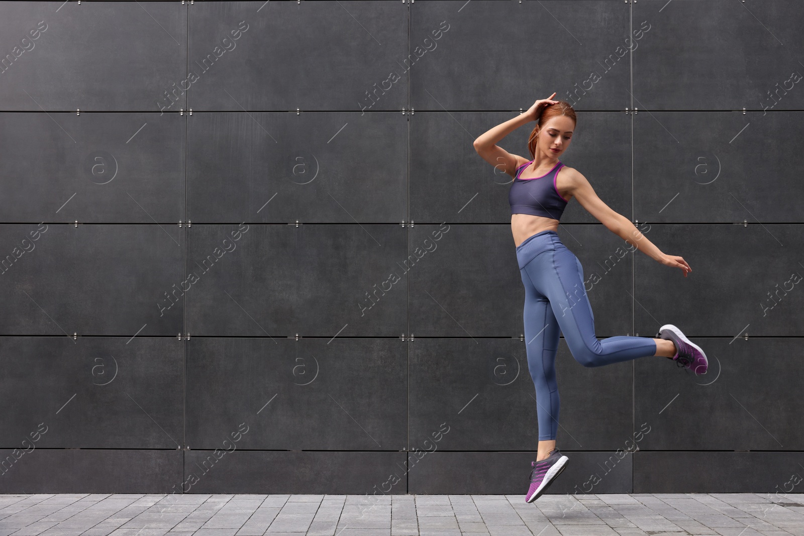 Photo of Beautiful woman in gym clothes jumping near dark grey wall on street, space for text