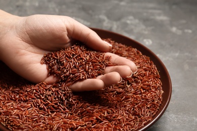 Photo of Woman holding grains near plate with brown rice on table, closeup