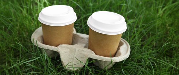 Takeaway paper coffee cups with plastic lids in cardboard holder on green grass outdoors