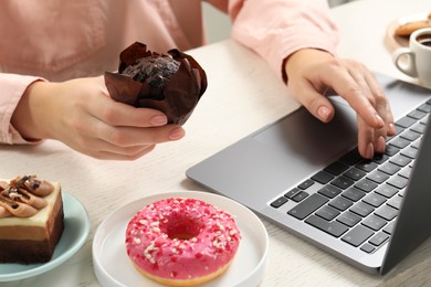 Photo of Bad habits. Woman eating muffin while using laptop at white wooden table, closeup