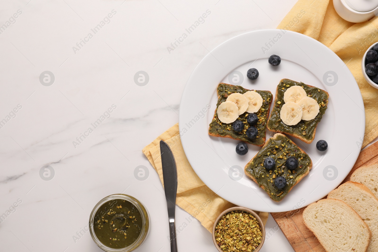 Photo of Toasts with tasty pistachio butter, banana, blueberries and nuts on white marble table, flat lay. Space for text