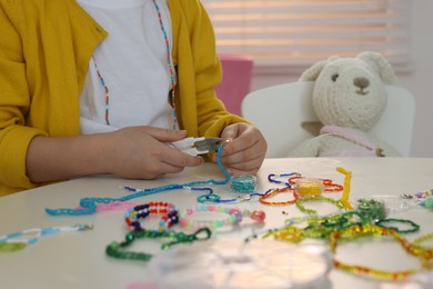 Photo of Little girl making beaded jewelry at table in room, closeup